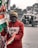 A person is holding several Indian flags in a street setting. They are wearing a red hoodie and a beige beanie, with a face mask covering their mouth. In the background, there is an auto-rickshaw with passengers and some buildings.
