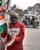 A person is holding several Indian flags in a street setting. They are wearing a red hoodie and a beige beanie, with a face mask covering their mouth. In the background, there is an auto-rickshaw with passengers and some buildings.