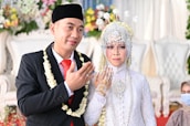 A newlywed couple dressed in traditional attire is posing for a photograph. The groom is wearing a black suit with a red tie, accompanied by a black traditional cap and a garland of flowers around his neck. The bride is adorned in an intricate white dress with detailed beadwork and a headdress with floral decorations. She has henna on her hand, and they are both showing their hands in a traditional gesture. The background features blurred floral decorations and a white upholstered chair.