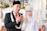 A newlywed couple dressed in traditional attire is posing for a photograph. The groom is wearing a black suit with a red tie, accompanied by a black traditional cap and a garland of flowers around his neck. The bride is adorned in an intricate white dress with detailed beadwork and a headdress with floral decorations. She has henna on her hand, and they are both showing their hands in a traditional gesture. The background features blurred floral decorations and a white upholstered chair.
