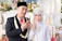 A newlywed couple dressed in traditional attire is posing for a photograph. The groom is wearing a black suit with a red tie, accompanied by a black traditional cap and a garland of flowers around his neck. The bride is adorned in an intricate white dress with detailed beadwork and a headdress with floral decorations. She has henna on her hand, and they are both showing their hands in a traditional gesture. The background features blurred floral decorations and a white upholstered chair.