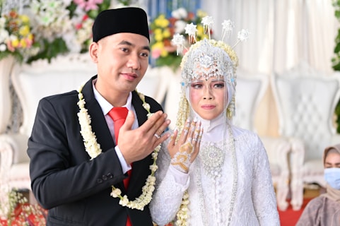 A newlywed couple dressed in traditional attire is posing for a photograph. The groom is wearing a black suit with a red tie, accompanied by a black traditional cap and a garland of flowers around his neck. The bride is adorned in an intricate white dress with detailed beadwork and a headdress with floral decorations. She has henna on her hand, and they are both showing their hands in a traditional gesture. The background features blurred floral decorations and a white upholstered chair.
