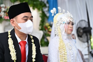 a man wearing a face mask sitting next to a woman in a wedding dress