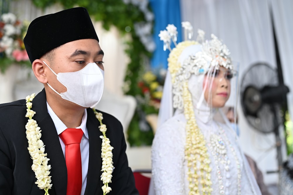 a man wearing a face mask sitting next to a woman in a wedding dress