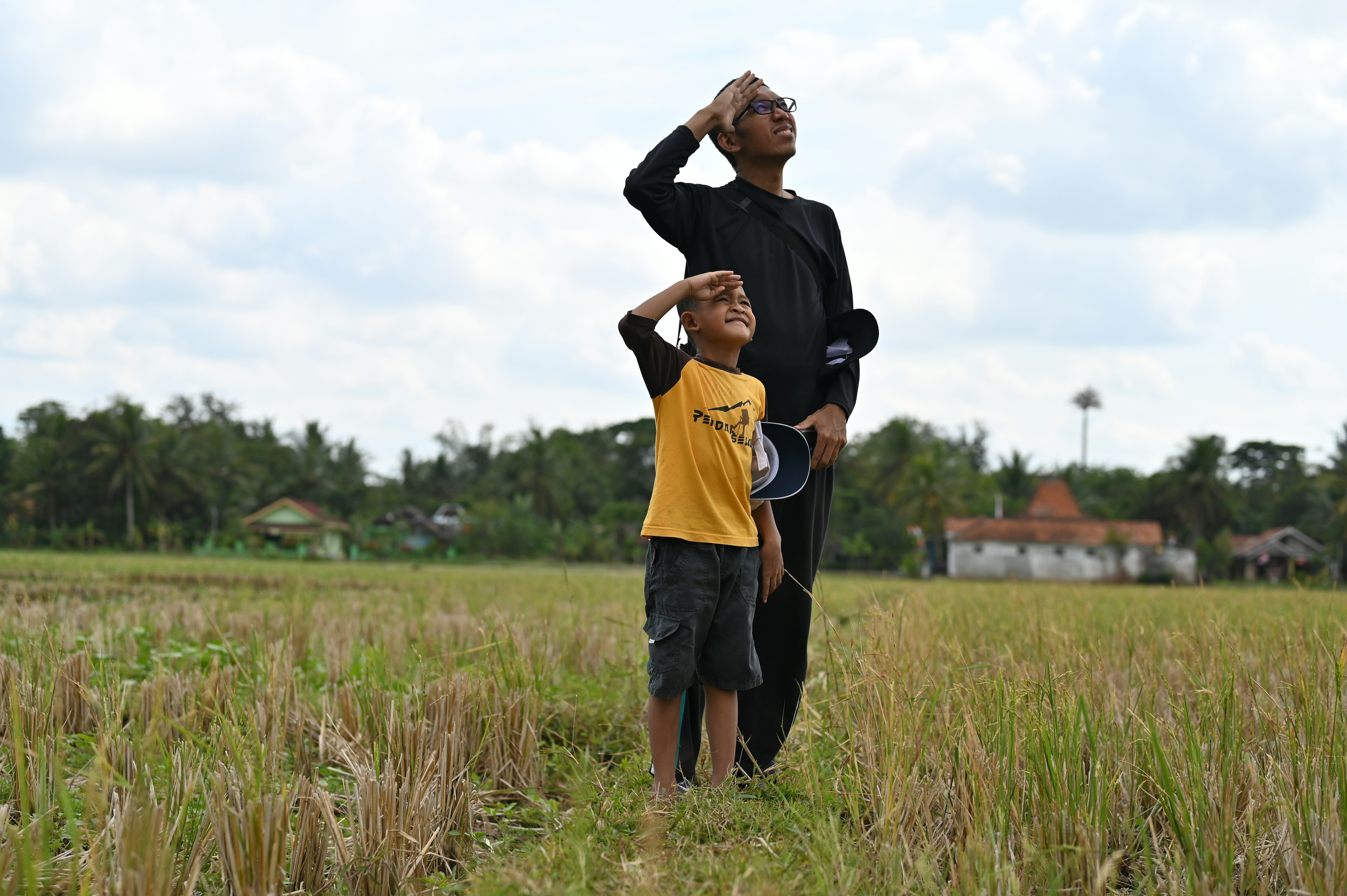 Two people stand in a golden field, shading their eyes as they gaze toward the cloudy sky.