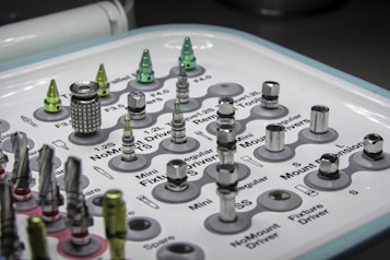 A tray containing various dental implants and tools organized in a grid. The metallic objects come in different shapes and sizes, some with shiny surfaces. Labels next to each object provide identification for their function or size. The overall setup appears clean and clinical, indicative of a medical or dental environment.