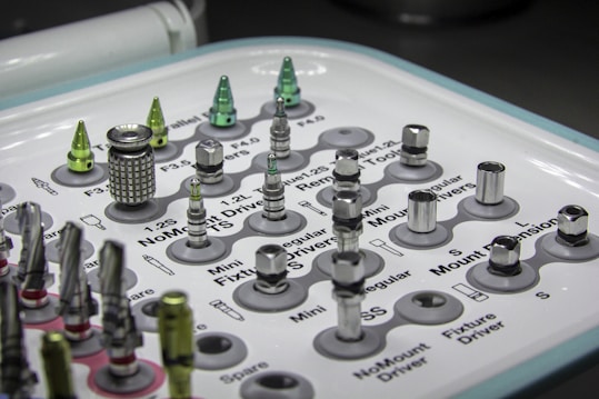 A tray containing various dental implants and tools organized in a grid. The metallic objects come in different shapes and sizes, some with shiny surfaces. Labels next to each object provide identification for their function or size. The overall setup appears clean and clinical, indicative of a medical or dental environment.