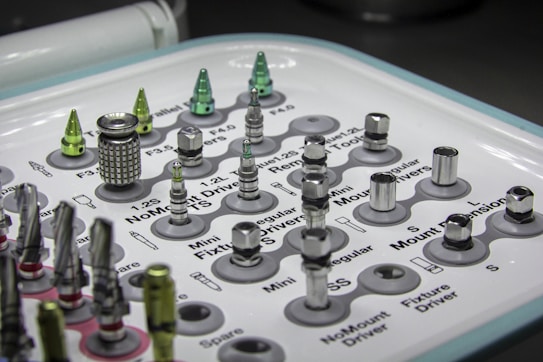 A tray containing various dental implants and tools organized in a grid. The metallic objects come in different shapes and sizes, some with shiny surfaces. Labels next to each object provide identification for their function or size. The overall setup appears clean and clinical, indicative of a medical or dental environment.