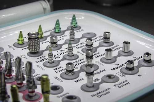 A tray containing various dental implants and tools organized in a grid. The metallic objects come in different shapes and sizes, some with shiny surfaces. Labels next to each object provide identification for their function or size. The overall setup appears clean and clinical, indicative of a medical or dental environment.