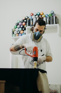 A person wearing a respirator mask and gloves operates an electric drill in a workspace. In the background, shelves contain neatly arranged spray paint cans of various colors.