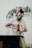 A person wearing a respirator mask and gloves operates an electric drill in a workspace. In the background, shelves contain neatly arranged spray paint cans of various colors.