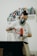 A person wearing a respirator mask and gloves operates an electric drill in a workspace. In the background, shelves contain neatly arranged spray paint cans of various colors.