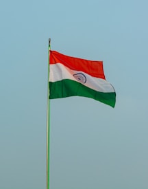 The image features a tricolor flag with horizontal bands of saffron, white, and green. A navy blue wheel with 24 spokes is centered in the white band. The flag is flying against a clear, pale blue sky.