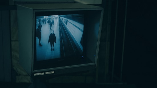 A surveillance monitor displays a live feed of a subway platform with a few people walking and standing. The monitor is enclosed in a box and situated in a dimly lit area, giving a sense of observation and monitoring in a transportation setting.