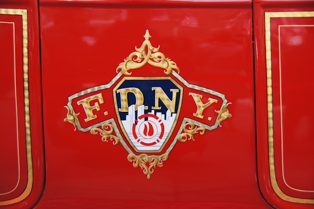 A red surface featuring a decorative emblem with 'FDNY' written on it, surrounded by gold embellishments and stylized lettering. The background includes a silhouette of a cityscape and a blue and gold color scheme.