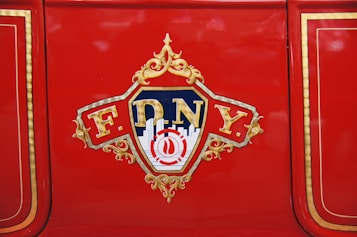 A red surface featuring a decorative emblem with 'FDNY' written on it, surrounded by gold embellishments and stylized lettering. The background includes a silhouette of a cityscape and a blue and gold color scheme.