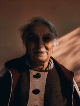 An elderly person with short gray hair is captured in warm lighting, casting soft shadows on their face. They wear a coat with a textured fabric featuring large buttons and seem to have a gentle, contemplative expression.