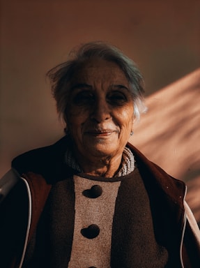 An elderly person with short gray hair is captured in warm lighting, casting soft shadows on their face. They wear a coat with a textured fabric featuring large buttons and seem to have a gentle, contemplative expression.