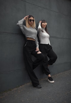 Two women lean casually against a dark gray wall. They are both wearing sunglasses and similarly styled outfits, including long-sleeved tops and black pants. One woman has a light gray cropped sweater, while the other wears a white sweater. The scene conveys a relaxed and stylish vibe.