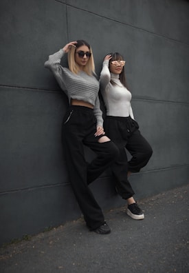 Two women lean casually against a dark gray wall. They are both wearing sunglasses and similarly styled outfits, including long-sleeved tops and black pants. One woman has a light gray cropped sweater, while the other wears a white sweater. The scene conveys a relaxed and stylish vibe.