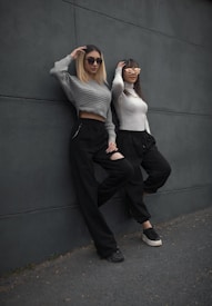 Two women lean casually against a dark gray wall. They are both wearing sunglasses and similarly styled outfits, including long-sleeved tops and black pants. One woman has a light gray cropped sweater, while the other wears a white sweater. The scene conveys a relaxed and stylish vibe.