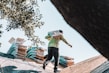 A person is carrying a bag of materials on a roof, with stacks of materials in the background. They are wearing a long-sleeve shirt and a cap. The scene includes tree branches partially framing the top left corner.
