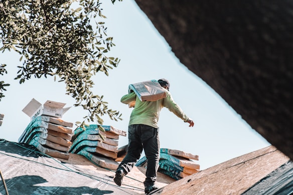 A person is carrying a bag of materials on a roof, with stacks of materials in the background. They are wearing a long-sleeve shirt and a cap. The scene includes tree branches partially framing the top left corner.
