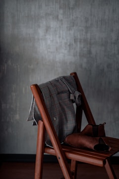A plaid shirt is draped over the back of a wooden chair, accompanied by a pair of brown leather boots placed on the seat. The background features a textured gray wall adding a rustic and homely feel to the scene.