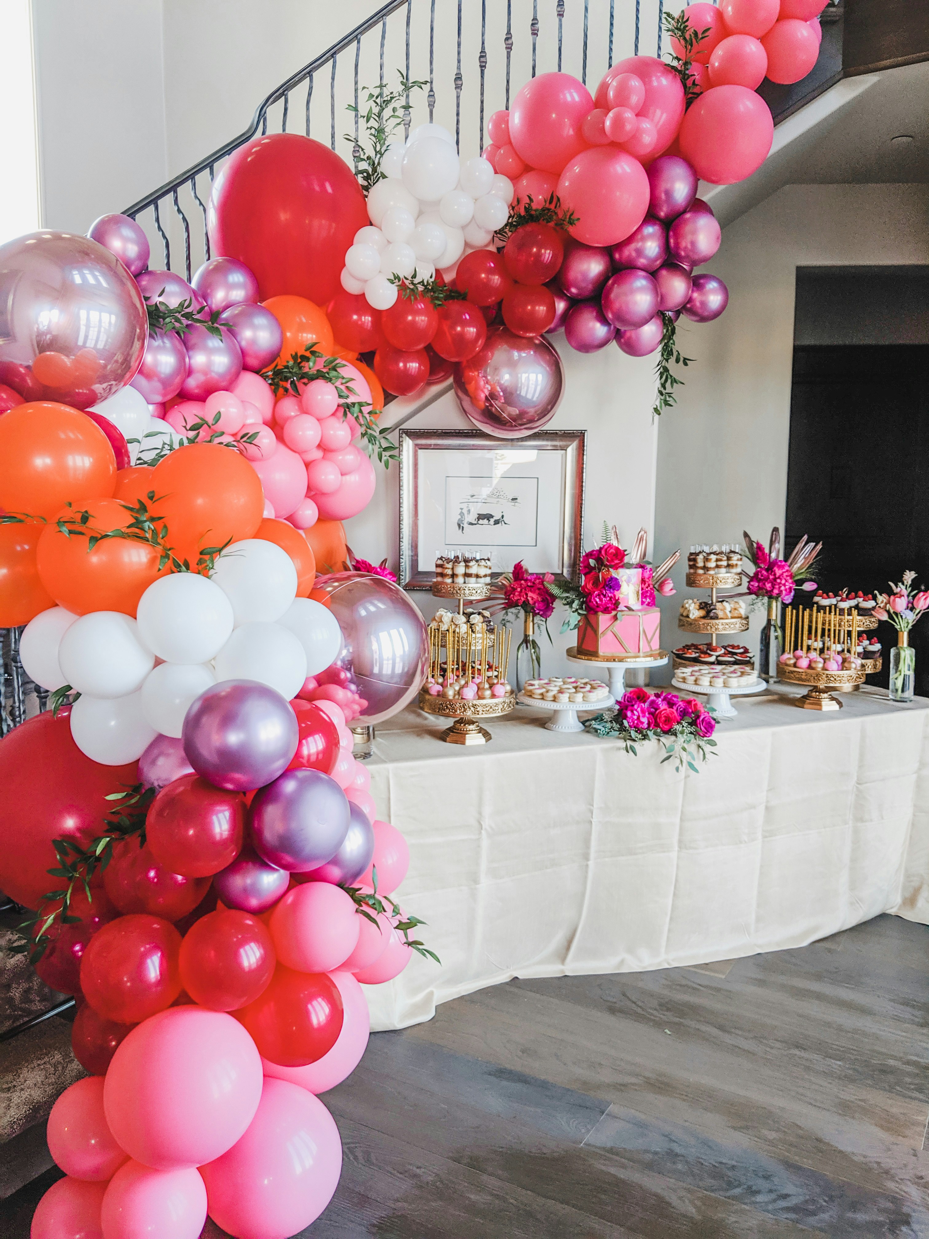 Festive birthday party setup with colorful balloon arch, decorated cake table, and fairy lights in a bright indoor venue