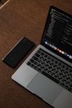 A laptop with an open screen displaying code is placed on a wooden table next to a smartphone. The laptop keyboard features Russian characters, and the screen shows a dark-themed development environment.