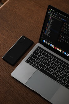 A laptop with an open screen displaying code is placed on a wooden table next to a smartphone. The laptop keyboard features Russian characters, and the screen shows a dark-themed development environment.