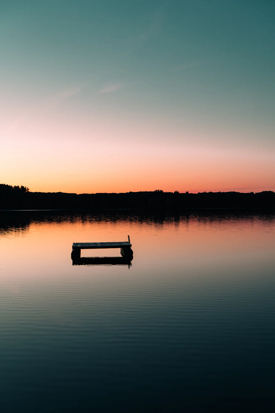 A floating swim dock on a calm lake at sunset, with a dark tree line on the far shore