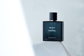 A minimalist composition featuring a dark, elegant bottle of Bleu de Chanel Eau de Parfum resting on a light surface. The background is soft and subtle, enhancing the focus on the sleek, rectangular bottle with its bold, white lettering.