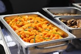 A stainless steel buffet tray is filled with a vibrant dish consisting of large chunks of bright orange squash and garnished with green beans. Surrounding trays contain different food items, adding to the variety of a buffet setup.