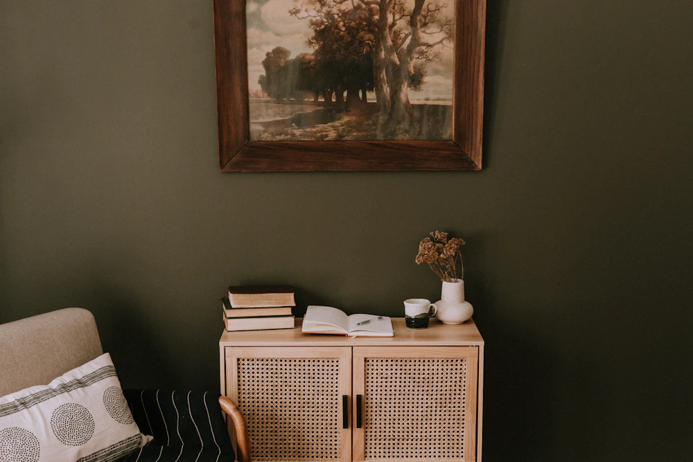 An olive green wall above a rattan cabinet with books and dried flowers