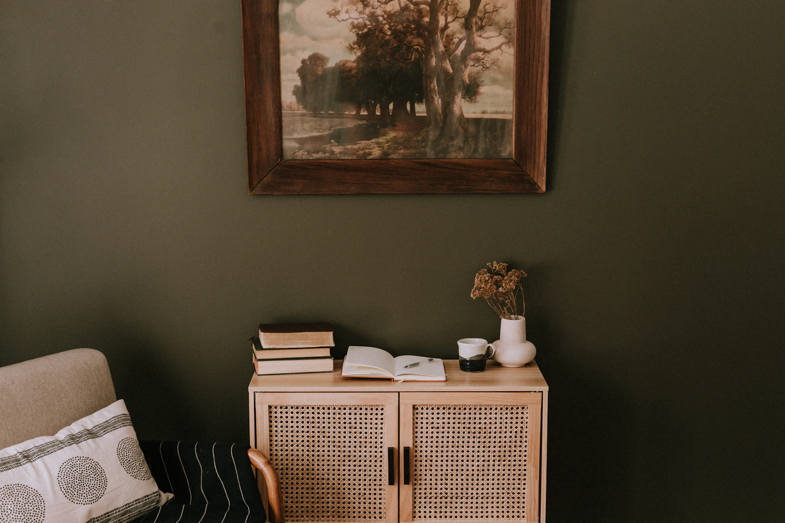 Deep olive wall with a landscape painting above a rattan-fronted cabinet holding books and dried flowers