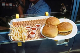A drive-in meal consisting of burgers and french fries is placed on a car's window-mounted tray. Three small cups of ketchup are arranged neatly beside the crispy fries. The burgers are served on a white paper plate, and there's a person visible inside the car, along with a small dog in the background.