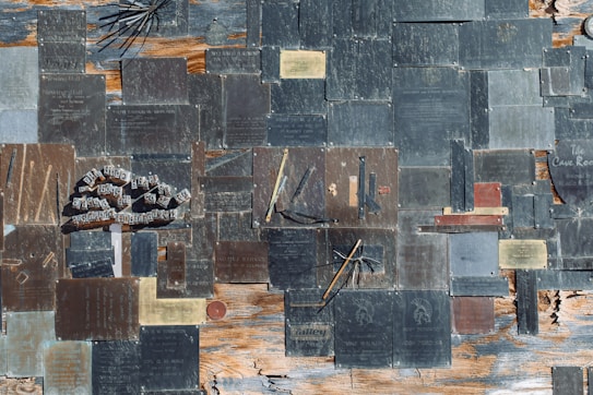 The image features an assortment of metal plaques mounted on a weathered wooden surface. The plaques are of various sizes and shapes, some with text inscriptions, while others have designs or symbols. The wood beneath shows signs of age, with peeling paint and visible grain. A collection of metal rods and wires, seemingly part of some installation, is interspersed among the plaques.
