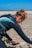 A person wearing a wetsuit is setting up equipment on a sandy beach. The individual has reddish-brown hair tied back and is holding kiteboarding lines. In the background, several colorful kites are visible in the sky against a clear blue backdrop.