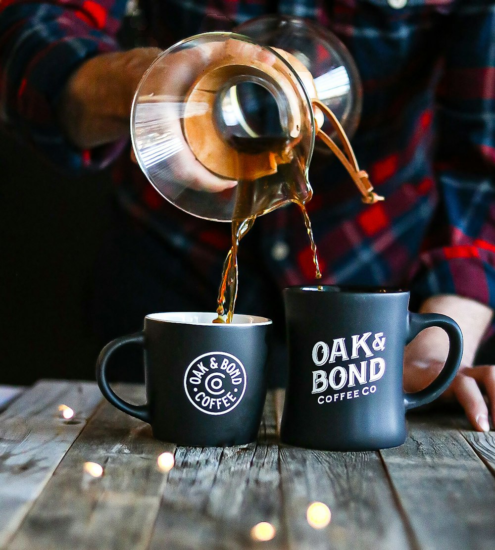 a person pouring coffee into two black coffee mugs