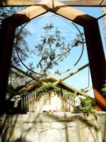 A chapel with a large circular window framing an intricate tree against a clear blue sky. The altar is adorned with green foliage, white flowers, and multiple candles on either side. An open book, likely a Bible, and a wooden cross are placed on the altar. The phrase 'OUR FATHER IN HEAVEN' is inscribed on the stone wall below the altar.