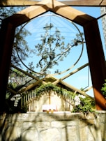 A chapel with a large circular window framing an intricate tree against a clear blue sky. The altar is adorned with green foliage, white flowers, and multiple candles on either side. An open book, likely a Bible, and a wooden cross are placed on the altar. The phrase 'OUR FATHER IN HEAVEN' is inscribed on the stone wall below the altar.