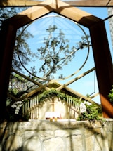 A chapel with a large circular window framing an intricate tree against a clear blue sky. The altar is adorned with green foliage, white flowers, and multiple candles on either side. An open book, likely a Bible, and a wooden cross are placed on the altar. The phrase 'OUR FATHER IN HEAVEN' is inscribed on the stone wall below the altar.