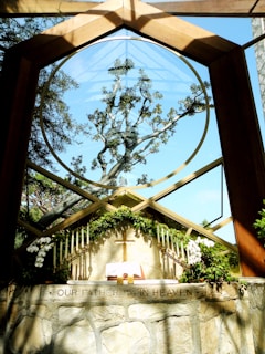 A chapel with a large circular window framing an intricate tree against a clear blue sky. The altar is adorned with green foliage, white flowers, and multiple candles on either side. An open book, likely a Bible, and a wooden cross are placed on the altar. The phrase 'OUR FATHER IN HEAVEN' is inscribed on the stone wall below the altar.