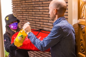 A delivery person wearing a purple mask, black hat with yellow designs, and black clothing is handing a large red insulated bag and a bottle of Jarritos pineapple soda to a bald man in a blue denim shirt at a door. The setting includes a brick wall and a carved wooden door.