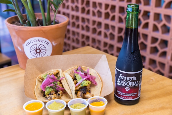 A wooden table holds a plate with two tacos garnished with minced meat, pickled red onions, and avocado sauce. Beside the plate are small containers of various sauces, and to the right is a cold bottle of Sangría Señorial. A potted plant against a patterned background is in the upper left corner.
