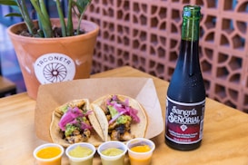 A wooden table holds a plate with two tacos garnished with minced meat, pickled red onions, and avocado sauce. Beside the plate are small containers of various sauces, and to the right is a cold bottle of Sangr&iacute;a Se&ntilde;orial. A potted plant against a patterned background is in the upper left corner.