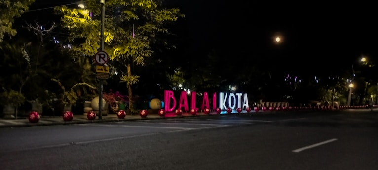 Illuminated large sign with the letters 'BALAI KOTA' on the roadside, surrounded by red spherical lights. Trees and a no parking sign are in the background, with the night sky and a partial view of the moon.