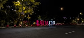 Illuminated large sign with the letters 'BALAI KOTA' on the roadside, surrounded by red spherical lights. Trees and a no parking sign are in the background, with the night sky and a partial view of the moon.