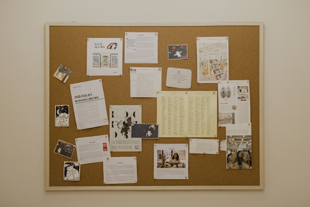 A cork bulletin board attached to a wall, filled with various documents, images, and flyers pinned. The contents include photos, printed text pages, graphic illustrations, and lists. Several sheets are loosely organized and thumbtacked to the board.